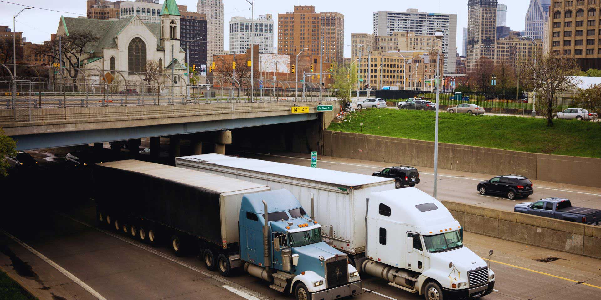 Two semi trucks traveling a highway with the city of Detroit in the background