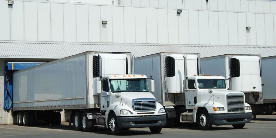 White semi trucks with reefer trailers backed up to the loading dock at a cold storage warehouse facility.