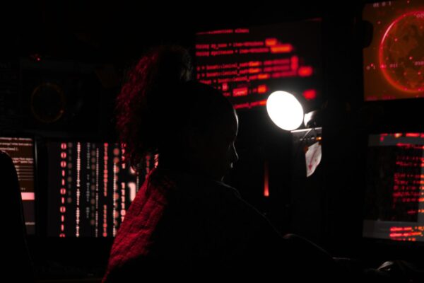 A silhouetted women working at a set of computer screens. She's engaged in freight fraud, empowered by generative AI tools.